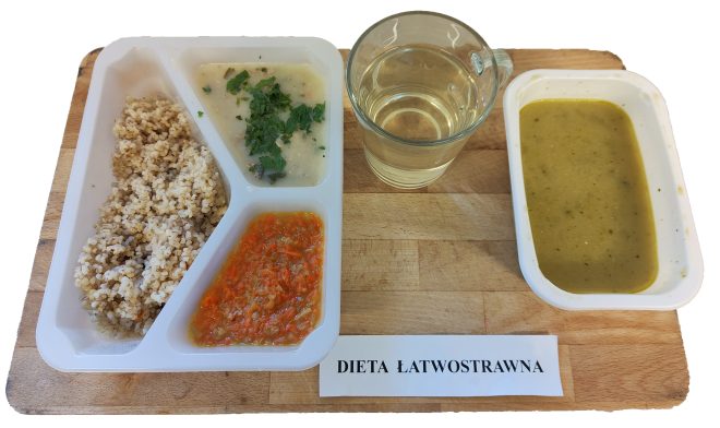 Pierwsze zdjęcie przedstawia zestaw obiadowy oznaczony jako „dieta łatwostrawna”, ułożony na jasnej, drewnianej tacy. W plastikowym pojemniku z trzema przegródkami znajdują się: porcja drobnoziarnistej kaszy (najpewniej jęczmiennej), jasny sos z dodatkiem świeżych ziół oraz warzywny dodatek z duszonej marchwi. Obok umieszczono szklankę z klarownym naparem lub kompotem oraz osobny pojemnik z gęstą, zielono-żółtą zupą kremem, prawdopodobnie warzywną. Całość została opisana etykietą „DIETA ŁATWOSTRAWNA”.