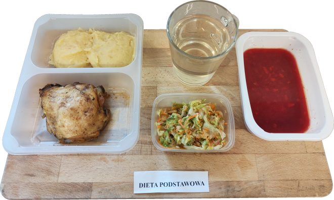 Posiłek obiadowy oznaczony etykietą „DIETA PODSTAWOWA”, podany na drewnianej tacy. Główne danie znajduje się w dwukomorowym pojemniku – z jednej strony umieszczono dwa kopiaste kleksy purée ziemniaczanego, z drugiej strony spory kawałek pieczonego kurczaka (prawdopodobnie udko lub ćwiartka bez panierki, z przypieczoną skórką). Obok, w osobnym pudełku, znajduje się surówka z białej kapusty z dodatkiem marchewki i koperku. Po prawej stronie stoi przezroczysty kubek ze zwykłą wodą oraz głęboki pojemnik z czerwoną zupą – wygląda na barszcz czerwony z drobną wkładką warzywną. Taca ustawiona na jasnym tle