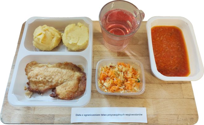 „Dieta z ograniczeniem łatwo przyswajalnych węglowodanów”. W plastikowym pojemniku widoczne są dwie porcje purée ziemniaczanego o kremowej konsystencji, uformowane w okrągłe kopce. Obok znajduje się panierowany kotlet schabowy o nieregularnym kształcie i złocistej, chrupiącej powierzchni. Dodatkowo, w osobnym przezroczystym pojemniczku podano surówkę z kiszonej kapusty z tartą marchewką oraz drobno posiekaną natką pietruszki – kolorowa mieszanka prezentuje się świeżo i apetycznie. Na tacy znajduje się także szklany kubek z różowym, przezroczystym napojem, prawdopodobnie kompotem owocowym lub herbatą malinową. Całość uzupełnia porcja zupy jarzynowej w oddzielnym plastikowym naczyniu – zupa ma pomidorową bazę i zawiera rozdrobnione warzywa, głównie marchew i seler
