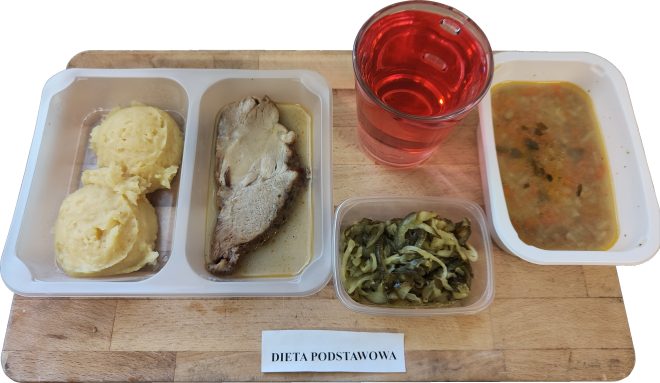 Obiad dla pacjenta na diecie podstawowej, podany na drewnianej tacy. W podłużnym pojemniku z przegródkami znajdują się dwie kulki puree ziemniaczanego oraz spory plaster pieczonego mięsa wieprzowego lub drobiowego w jasnym sosie pieczeniowym. Obok, w oddzielnym pojemniczku – gotowana kapusta z dodatkiem przypraw. W zestawie znajduje się również porcja zupy jarzynowej z wyczuwalnymi kawałkami marchwi, ziemniaków i ziół, a także przezroczysta szklanka z intensywnie czerwoną herbatą owocową lub naparem. Etykieta wskazuje, że to „DIETA PODSTAWOWA”