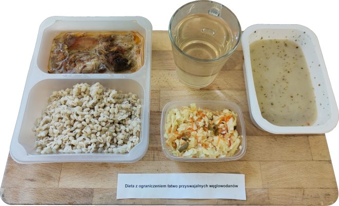 Obiad serwowany w pojemniku z trzema przegródkami i dodatkowymi naczyniami, opisany jako „Dieta z ograniczeniem łatwo przyswajalnych węglowodanów”. W głównych przegródkach znajdują się: gotowany jęczmień, duszone mięso z warzywami w galarecie oraz szklanka przezroczystego kompotu lub naparu. W osobnym pojemniczku podano surówkę z białej kapusty z marchewką i ogórkiem kiszonym. Zestaw uzupełnia miseczka z gęstą, jasną zupą warzywną. Wszystko umieszczone na jasnej desce