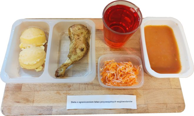 Zestaw obiadowy ułożony na drewnianej desce. Po lewej stronie tacki znajduje się porcja purée ziemniaczanego w postaci trzech kulek, obok leży pieczone udko kurczaka przyprawione ziołami. Zestaw uzupełnia mały pojemnik z surówką z marchwi i selera, miseczka z zupą o gładkiej, pomarańczowej konsystencji oraz szklanka czerwonego kompotu. Pod zestawem widoczna etykieta z napisem: „Dieta z ograniczeniem łatwo przyswajalnych węglowodanów”
