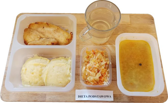 Zestaw obiadowy w pojemniku typu cateringowego z podpisem „Dieta podstawowa”. W skład dania wchodzi panierowany filet z kurczaka lub indyka, dwie porcje ziemniaczanego purée z koperkiem oraz surówka z kapusty kiszonej z marchwią. Obok – porcja zupy (najpewniej jarzynowej lub ziemniaczanej) o żółtawym zabarwieniu z widoczną tłuszczową okrągłą plamką oraz przezroczysty kubek z napojem, prawdopodobnie kompotem. Wszystko ułożone na drewnianej tacy