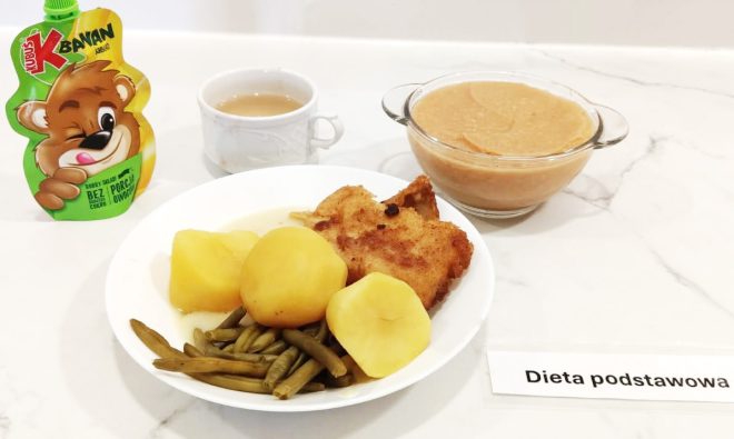Na pierwszym zdjęciu widoczny jest posiłek obiadowy składający się z gotowanych ziemniaków, panierowanego i smażonego kotleta (najprawdopodobniej drobiowego), oraz porcji gotowanej zielonej fasolki szparagowej. Obok dania głównego znajduje się filiżanka z jasnym naparem herbaty oraz miseczka pełna gęstego, jasnobrązowego kompotu z owoców. Dodatkowo, po lewej stronie umieszczono owocowy mus jabłkowy dla dzieci w saszetce marki Kubuś. Posiłek został opisany etykietą „Dieta podstawowa” i podany na białym tle kuchennego blatu