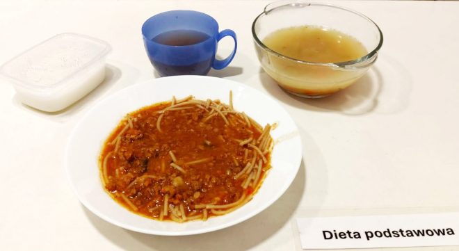 Drugie zdjęcie przedstawia obiad złożony z makaronu typu spaghetti w gęstym sosie bolońskim z mielonym mięsem i warzywami. Danie podano na dużym białym talerzu. Towarzyszy mu miseczka z klarownym, złotobrązowym bulionem oraz plastikowy kubek z ciemną herbatą. W tle znajduje się przezroczysty pojemnik z białym dodatkiem – prawdopodobnie deserem mlecznym lub jogurtem naturalnym. Wszystko ułożone jest na jasnym blacie z podpisem „Dieta podstawowa”
