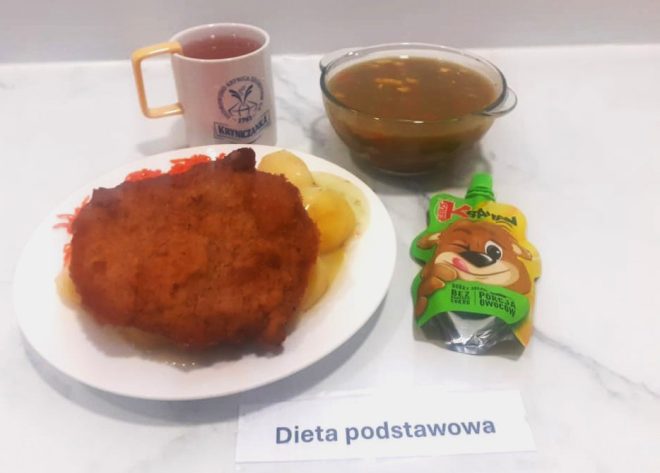 Na zdjęciu widoczny jest zestaw obiadowy w ramach diety podstawowej, ułożony na jasnym, marmurowym blacie. Na talerzu znajduje się panierowany kotlet schabowy, podany z gotowanymi ziemniakami i surówką z marchewki. Obok talerza ustawiono kubek z herbatą z logo Zakładu Opiekuńczo-Leczniczego „Kryniczanka”, szklane naczynie z gorącą zupą (prawdopodobnie jarzynową) oraz mus owocowy w tubce dla dzieci marki Kubuś. Przed zestawem umieszczono pasek z napisem „Dieta podstawowa”