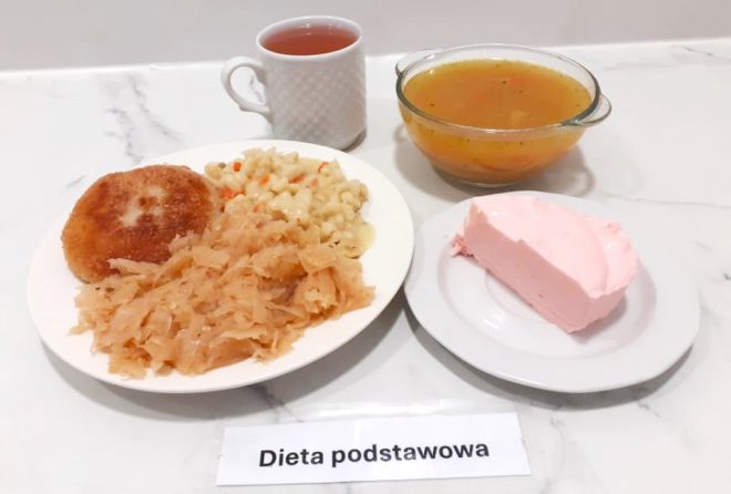 Obiad w wersji podstawowej sfotografowany na marmurowym blacie. Na talerzu znajduje się panierowany kotlet mielony, duża porcja gotowanej kapusty oraz makaron z warzywami. W tle stoi kubek z herbatą oraz miska z zupą warzywną na wywarze. Dodatkowo podano deser – różowy mus lub piankę na białym talerzyku. Na pierwszym planie widnieje kartka z napisem „Dieta podstawowa”