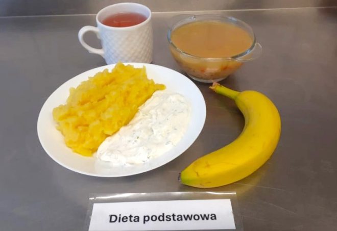 Obiad w diecie podstawowej podany na metalicznym blacie. Na talerzu po lewej stronie znajduje się porcja ziemniaków purée z dodatkiem marchewki oraz biały sos koperkowy lub tzatziki. Obok umieszczono całego banana, przezroczystą miseczkę z klarowną zupą jarzynową z ryżem oraz kubek z herbatą. Przed zestawem widnieje karteczka z etykietą „Dieta podstawowa”. Kompozycja posiłku sugeruje lekkie i pełnowartościowe danie obiadowe