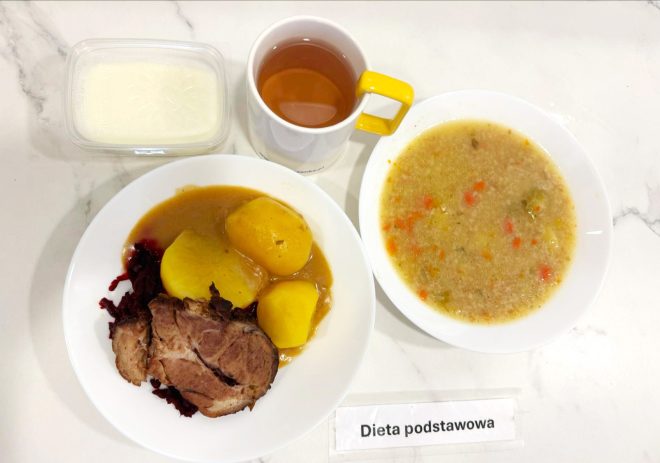 Zdjęcie przedstawia obiad z diety podstawowej. Po lewej stronie widoczny jest talerz z dwoma ziemniakami polanymi sosem pieczeniowym, porcją duszonych buraczków oraz kawałkiem pieczonego mięsa (najprawdopodobniej karkówka). Po prawej stronie znajduje się talerz z gęstą zupą jarzynową na kaszy jęczmiennej. W tle stoi przezroczyste pudełeczko z białym deserem mlecznym (możliwe, że budyń) oraz kubek z przezroczystą herbatą w białym kubku z żółtym uchem. Posiłek oznaczony został karteczką z napisem „Dieta podstawowa”