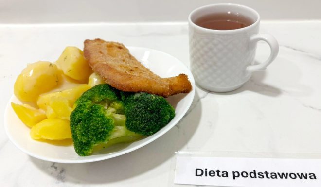 Na białym talerzu ułożono porcję obiadową w ramach diety podstawowej. Na pierwszym planie widoczne są ugotowane na parze lub w wodzie różyczki brokułów, zachowujące intensywnie zielony kolor. Obok znajdują się złociste ziemniaki w całości, polane lekkim sosem koperkowym. W górnej części talerza leży panierowany i smażony filet rybny o chrupiącej, rumianej skórce. Całość podana została na białym blacie, a obok ustawiono biały porcelanowy kubek z gorącym naparem – prawdopodobnie z herbatą. Przed talerzem umieszczono podpis z nazwą posiłku: „Dieta podstawowa”