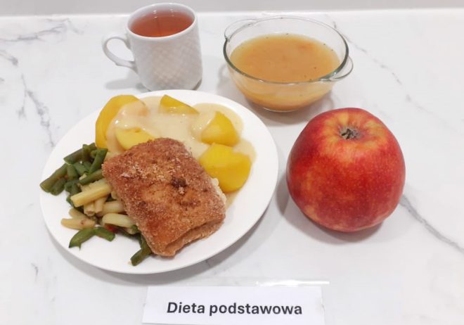 Zestaw obiadowy podany na białym tle, oznaczony jako „Dieta podstawowa”. Na talerzu znajduje się panierowany filet rybny, ugotowane ziemniaki polane jasnym sosem oraz porcja warzyw gotowanych – fasolki szparagowej zielonej i żółtej. W tle ustawiono filiżankę z gorącym napojem (najpewniej herbata) oraz miseczkę z zupą – o klarownej, bursztynowej barwie z widocznymi drobinkami warzyw. Całość uzupełnia duże czerwone jabłko jako element owocowy posiłku