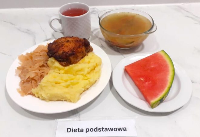 Na marmurowym blacie rozłożony jest pełnowartościowy obiad zgodny z dietą podstawową. Na głównym talerzu widać porcję purée ziemniaczanego, duszoną kapustę kiszoną oraz solidny kawałek pieczonego udka z kurczaka o rumianej skórce. Z boku znajduje się głęboki talerz z klarowną zupą – prawdopodobnie jarzynową – serwowaną w przezroczystej miseczce. Dodatkowo podany jest plaster soczystego arbuza na osobnym białym talerzyku. Obok posiłku stoi biały kubek z herbatą owocową lub kompotem. Pod talerzami widnieje etykieta z podpisem „Dieta podstawowa”
