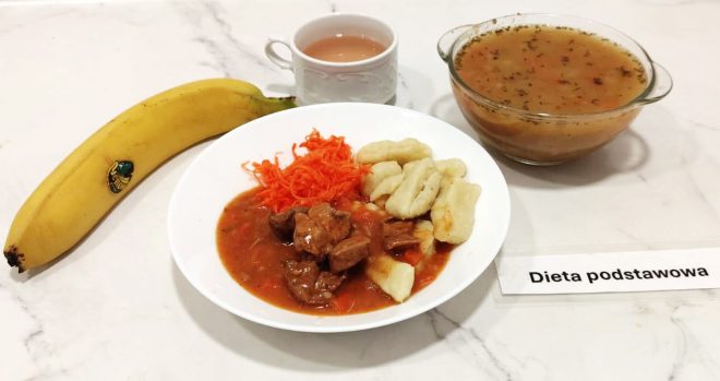 Obiad w ramach diety podstawowej ułożony na marmurowym blacie. Na dużym, białym talerzu znajduje się porcja gulaszu wołowego z sosem, obok kopytka i tarta marchewka. Po lewej stronie talerza leży dojrzały banan. W tle znajduje się filiżanka z herbatą z mlekiem oraz szklana miska z klarowną zupą – prawdopodobnie rosołem – z widocznymi drobinkami ziół. Przed zestawem umieszczona została etykieta z napisem „Dieta podstawowa”. Całość prezentuje się jako sycący i klasyczny posiłek obiadowy, przeznaczony dla pacjentów bez specjalnych ograniczeń dietetycznych