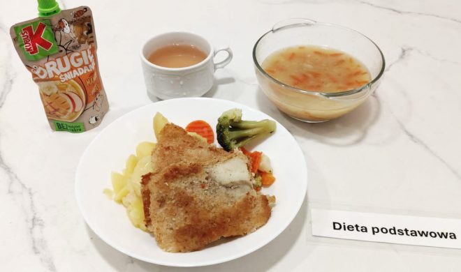 Obiad serwowany na białym marmurowym blacie z widoczną etykietą „Dieta podstawowa”. Na głównym talerzu znajduje się panierowany filet rybny, gotowane ziemniaki oraz gotowane warzywa: marchew, brokuł i kalafior. Obok ustawiona jest przezroczysta miska z zupą jarzynową, w której widoczne są kawałki marchewki i ziemniaków w delikatnym wywarze. Zestaw uzupełnia biały kubek z herbatą z mlekiem oraz owocowy mus Kubuś „Drugie śniadanie” o smaku jabłkowo-marchewkowym w saszetce typu „do wyciskania”