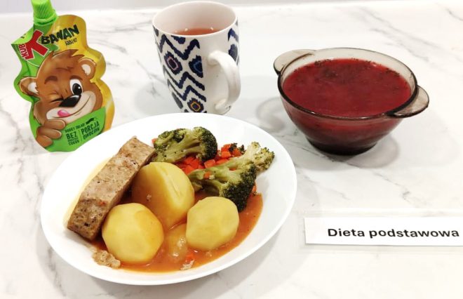 Na białym talerzu znajduje się porcja obiadowa składająca się z trzech gotowanych ziemniaków, plasterka pieczeni w sosie, gotowanych brokułów i marchewki. Obok talerza stoi kubek z napojem owocowym, szklana miseczka z czerwonym barszczem oraz mus owocowy w saszetce dla dzieci. W prawym dolnym rogu widoczna jest etykieta z napisem „Dieta podstawowa”. Całość sfotografowana na jasnym, marmurowym blacie