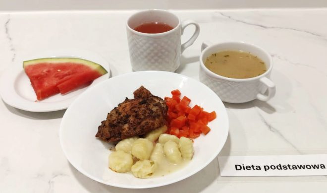 Na białym tle, na zestawie obiadowym oznaczonym jako „Dieta podstawowa”, podano posiłek złożony z kilku elementów. Na głównym talerzu znajdują się: grillowany lub pieczony kawałek mięsa (najprawdopodobniej udko lub filet z kurczaka), kluski gnocchi polane sosem oraz gotowana marchewka pokrojona w drobną kostkę. Obok – miseczka z klarownym rosołem lub bulionem ziołowym oraz filiżanka z napojem owocowym. Dodatkowo na osobnym spodeczku znajduje się trójkątny plaster arbuza. Całość serwowana jest w eleganckiej, porcelanowej zastawie, na marmurowym blacie