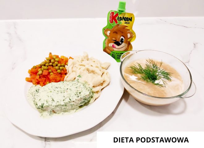 Na zdjęciu widoczny jest zestaw obiadowy zgodny z zasadami diety podstawowej, serwowany na białym tle kuchennym. Na dużym, białym talerzu znajduje się porcja gotowanego fileta rybnego w delikatnym sosie koperkowym, ułożonego z przodu po lewej stronie. Obok, po prawej stronie, znajduje się kopczyk jasnych klusek kładzionych, a nad nimi – mieszanka gotowanej marchewki i zielonego groszku. W tle, tuż za talerzem, stoi saszetka z musem jabłkowym marki Kubuś w wersji „Kawan jabłko”, ozdobiona rysunkiem uśmiechniętego misia. Po prawej stronie kadru ustawiono przezroczystą szklaną miseczkę z zupą – najprawdopodobniej kremem warzywnym – udekorowaną kleksem śmietany i świeżym koperkiem na wierzchu. W prawym dolnym rogu zdjęcia widnieje podpis: „DIETA PODSTAWOWA”