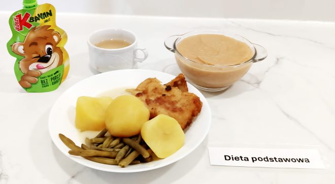 Na talerzu głównym: panierowany filet z ryby, trzy sztuki ziemniaków i gotowana fasolka szparagowa. Do zestawu podano przecier owocowy w szklanej miseczce, herbatę w białej filiżance i mus owocowy w saszetce dla dzieci (marki Lubus, banan-jabłko). Obok etykieta z napisem „Dieta podstawowa”