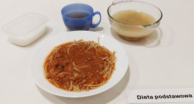 Na drugim zdjęciu zaprezentowano posiłek obiadowy. W centrum znajduje się biały talerz z porcją makaronu spaghetti w gęstym sosie bolońskim z mięsem mielonym i drobno pokrojonymi warzywami. Towarzyszą mu trzy dodatki: przezroczysta miska z klarownym bulionem, niebieski plastikowy kubek z ciemną herbatą oraz biały plastikowy pojemniczek z mlecznym deserem lub jogurtem naturalnym. Wszystko ułożono na jasnym blacie stołu z widoczną etykietą „Dieta podstawowa”