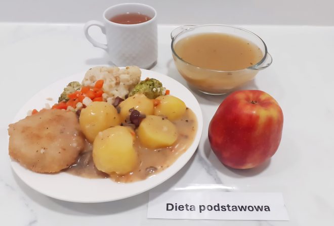 Zestaw obiadowy w ramach diety podstawowej, podany na białej tacy. Na talerzu ziemniaki z sosem i kawałkami pieczeni lub kotleta, gotowane warzywa (marchew, kalafior, brokuły) oraz duszony sos grzybowy. Obok miseczka z czystym bulionem, czerwone jabłko i filiżanka z naparem ziołowym lub kompotem. Posiłek wygląda pełnowartościowo i apetycznie, typowy dla klasycznego jadłospisu szpitalnego