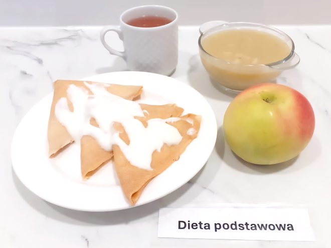 Na zdjęciu zaprezentowano zestaw śniadaniowy ustawiony na marmurowym blacie. W centrum znajduje się biały talerz z kilkoma plastrami sera żółtego ułożonymi w wachlarz, połówkami ugotowanego na twardo jajka, plastrem pomidora, liściem sałaty oraz porcją masła. Obok, po prawej stronie, na osobnym talerzyku leżą trzy kromki jasnego chleba pszennego. W tle widoczny jest ceramiczny kubek z logo „Kryniczanka”, wypełniony jasnym napojem – najpewniej herbatą z mlekiem lub kawą z mlekiem. Poniżej talerza znajduje się kartka z napisem „Dieta podstawowa”, wskazująca na rodzaj zastosowanego jadłospisu