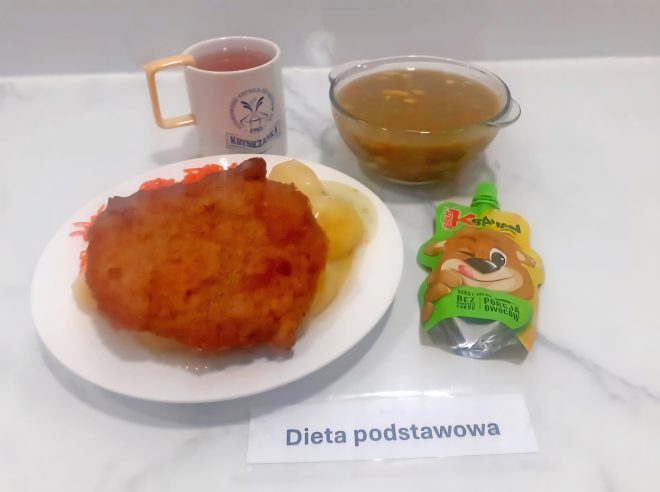 Na jasnym marmurowym blacie widoczna jest taca z podpisem „Dieta podstawowa”. Posiłek obiadowy składa się z kilku elementów. Na białym talerzu znajduje się panierowany, smażony kotlet schabowy podany z dwoma gotowanymi ziemniakami oraz startą surówką z marchewki. Kotlet i ziemniaki są polane lekkim sosem. Obok talerza ustawiono przezroczystą miskę z gorącą, klarowną zupą – najprawdopodobniej warzywną, z widocznymi cząstkami warzyw. W tle po lewej stronie widoczny jest kubek z herbatą z nadrukiem ośrodka „Kryniczanka”. Po prawej stronie ustawiono saszetkę owocowego musu w opakowaniu z wizerunkiem misia i napisem „Kubuś” – produkt bez dodatku cukru. Kompozycja posiłku wskazuje na klasyczny, pełnowartościowy obiad dostosowany do standardowej diety szpitalnej