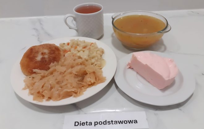 Na białym, marmurowym blacie znajduje się zestaw obiadowy opisany jako „Dieta podstawowa”. W centrum zdjęcia stoi talerz z czterema elementami: panierowanym kotletem (najprawdopodobniej mielonym), gotowaną kapustą kiszoną, drobnym makaronem z dodatkiem marchwi i sosem, który go oblepia. Po prawej stronie znajduje się szklana miska z przezroczystym rosołem z warzywami. W prawym dolnym rogu na osobnym talerzyku podano różowy deser w formie pianki lub galaretki mlecznej. Obok deseru stoi biały kubek z herbatą owocową lub ziołową. Całość wygląda schludnie, dania są porcjowane i podane w oddzielnych naczyniach