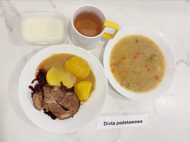 Na białym talerzu podano porcję obiadową w ramach diety podstawowej. Składa się ona z dwóch grubych plastrów pieczeni wieprzowej, dwóch gotowanych ziemniaków oraz buraczków na ciepło. Całość oblana jest jasnym sosem pieczeniowym. Obok, w białej misce, znajduje się talerz gęstej zupy jarzynowej z wyczuwalnymi kawałkami marchwi, ziemniaków i pora. Do posiłku podano herbatę w białym kubku z żółtym uchem oraz deser mleczny w plastikowym pojemniczku z przezroczystą pokrywką. Wszystko ułożono na marmurowym blacie, obok etykiety z napisem „Dieta podstawowa”