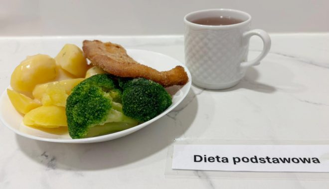 Na białym talerzu umieszczonym na marmurowym blacie podano zestaw obiadowy zgodny z dietą podstawową. Na pierwszym planie widoczne są ugotowane ziemniaki, polane lekkim sosem koperkowym, obok znajdują się intensywnie zielone różyczki brokułów. Na warzywach i ziemniakach spoczywa rumiany filet rybny w chrupiącej panierce, lekko zagięty na końcach. Po prawej stronie talerza ustawiono biały kubek z subtelnym wzorem rombów, wypełniony jasnym naparem – prawdopodobnie herbatą. W prawym dolnym rogu zdjęcia, na blacie, widoczna jest etykieta z czarnym napisem „Dieta podstawowa” umieszczona w przezroczystej folii. W tle kafelki ścienne w jasnym kolorze nadają całości estetyczny, sterylny wygląd