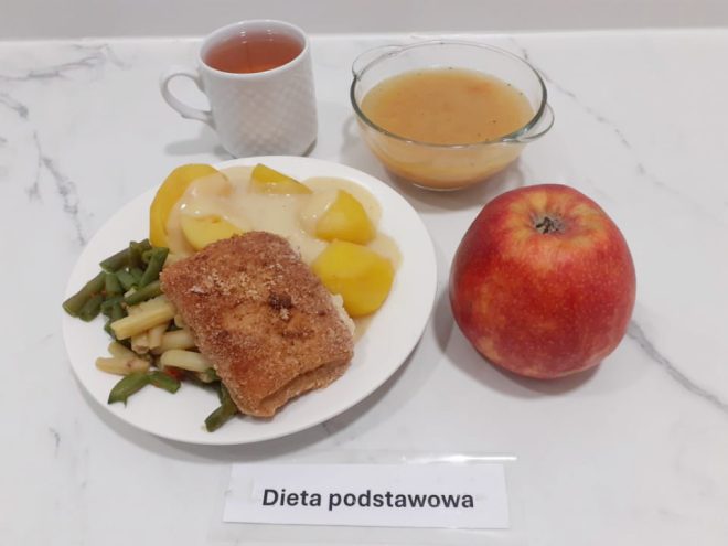 Na marmurowym blacie znajduje się talerz obiadowy z zestawem posiłku w diecie podstawowej. Na talerzu podano panierowany filet rybny, gotowane ziemniaki polane sosem oraz mieszankę warzyw z fasolki zielonej i żółtej. Obok znajduje się duże, czerwone jabłko oraz przezroczysta miseczka z klarowną zupą jarzynową o delikatnym zabarwieniu. Całość uzupełnia biały kubek z uchwytem, wypełniony herbatą. Przed posiłkiem umieszczono etykietę z napisem „Dieta podstawowa”