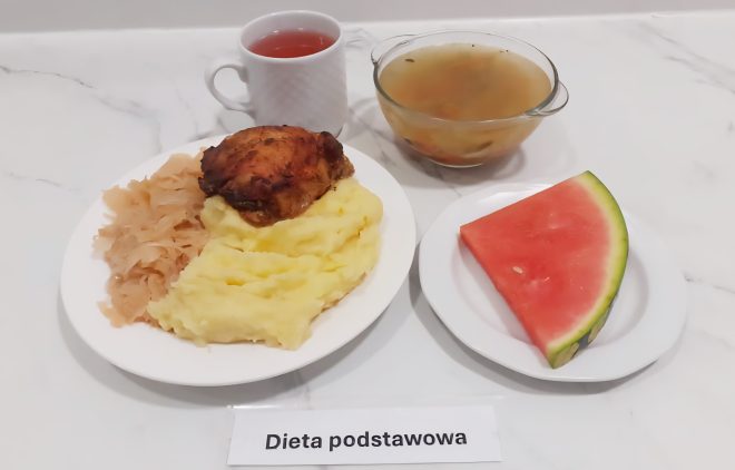 Pierwsze zdjęcie przedstawia pełnowartościowy obiad skomponowany w ramach diety podstawowej, podany na jasnym blacie. Na dużym talerzu znajduje się porcja puszystego purée ziemniaczanego, duszona kapusta oraz solidny kawałek pieczonego kurczaka z przyrumienioną skórką. Obok ustawiono miseczkę z klarowną zupą, prawdopodobnie rosołem z dodatkiem drobnych warzyw oraz filiżankę z herbatą owocową. Uzupełnieniem posiłku jest deser w postaci soczystego trójkątnego kawałka arbuza, ułożonego na osobnym białym talerzyku. Przed potrawami widnieje kartka z napisem „Dieta podstawowa”