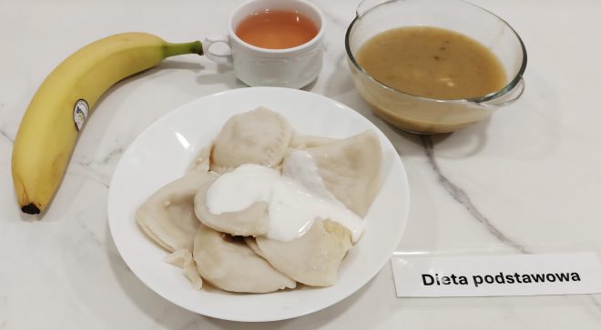 Zestaw obiadowy na diecie podstawowej, serwowany na marmurowym blacie. W centrum znajduje się biały głęboki talerz z pierogami (najprawdopodobniej ruskimi), polanymi śmietaną. Po lewej stronie widoczny cały banan oraz biały porcelanowy kubek z herbatą. Po prawej – szklana miska z gęstą zupą lub sosem, w kolorze jasnobrązowym. Przed talerzami – etykieta z napisem „Dieta podstawowa”