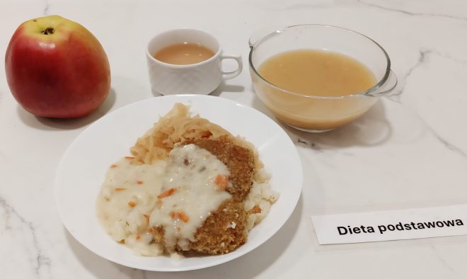 Zestaw obiadowy na białym marmurowym blacie składa się z kilku elementów: na dużym talerzu znajduje się panierowany kotlet drobiowy polany jasnym sosem warzywnym, porcja puree ziemniaczanego oraz duszona kiszona kapusta. Obok talerza postawiono dużą szklankę z przezroczystą zupą – prawdopodobnie kompotem owocowym – oraz białą filiżankę z napojem (prawdopodobnie herbata zbożowa). Całość uzupełnia świeże, czerwono-żółte jabłko. Z boku widoczna jest etykieta z napisem „Dieta podstawowa”