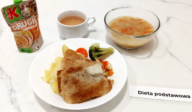 Na białym marmurowym blacie znajduje się zestaw obiadowy z etykietą „Dieta podstawowa”. Na talerzu umieszczono porcję gotowanych ziemniaków, warzywa na parze (brokuł, marchewka i kalafior) oraz panierowany filet rybny. Obok talerza znajduje się przezroczysta miska z zupą jarzynową z widocznymi paskami marchewki i ziemniakami. Dodatkowo podano biały kubek z herbatą z mlekiem oraz saszetkę owocowego musiku Kubuś „Drugie śniadanie” o smaku jabłkowo-marchewkowym bez dodatku cukru