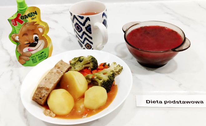 Na jasnym, marmurowym blacie ustawiono posiłek obiadowy w ramach diety podstawowej. Główny talerz zawiera trzy gotowane ziemniaki, plaster pieczeni wieprzowej w sosie, brokuły oraz gotowaną marchewkę pokrojoną w drobną kostkę. Obok umieszczono szklaną miseczkę z gorącym barszczem czerwonym oraz kubek z kolorowym wzorem, prawdopodobnie zawierający owocową herbatę. Po lewej stronie znajduje się mus owocowy w saszetce marki Kubuś, o smaku jabłkowym, bez dodatku cukru. Poniżej talerza widnieje etykieta z napisem „Dieta podstawowa”. Kompozycja posiłku odpowiada wymaganiom żywieniowym osób bez specjalnych ograniczeń dietetycznych