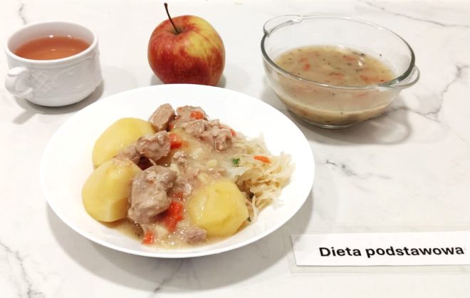 Na dużym białym talerzu podano porcję ziemniaków z gulaszem z mięsa wieprzowego i marchewką, a także dodatkiem kiszonej kapusty. Obok znajduje się przezroczysta miseczka z zupą jarzynową na bazie wywaru warzywnego oraz czerwone jabłko. W tle biały kubek z herbatą. Wszystko ustawione na jasnym blacie, z etykietą „Dieta podstawowa”