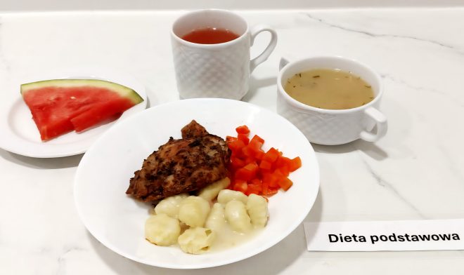 Zestaw obiadowy sfotografowany na marmurowym blacie, opatrzony etykietą „Dieta podstawowa”. W centrum znajduje się głęboki talerz z daniem głównym: pieczony kawałek mięsa (prawdopodobnie udko kurczaka bez kości) ułożony na porcję klusek gnocchi, polanych jasnym sosem. Obok podano gotowaną marchewkę pokrojoną w drobną kostkę. Do posiłku dołączono filiżankę z czerwonawym naparem (najpewniej herbata owocowa) oraz bulion z ziołami, serwowany w porcelanowej miseczce z uszkami. Na osobnym spodeczku leży trójkątny plaster arbuza. Za kadrem, ale przy zestawie – obecna jest osoba (autorka fotografii), dokumentująca posiłek