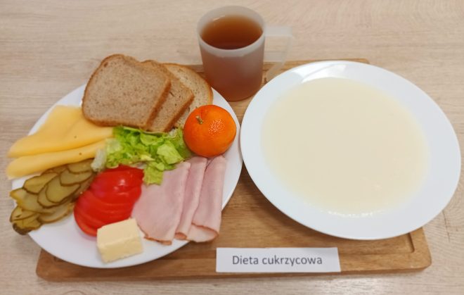 Na dużym talerzu znajduje się śniadaniowy zestaw: trzy kromki chleba razowego, plastry żółtego sera i szynki, sałata, pomidor i ogórek kiszony, kostka masła oraz mandarynka. Obok postawiono kubek z herbatą oraz głęboki talerz z jasną, kremową zupą mleczną. Posiłek oznaczony etykietą „Dieta cukrzycowa”