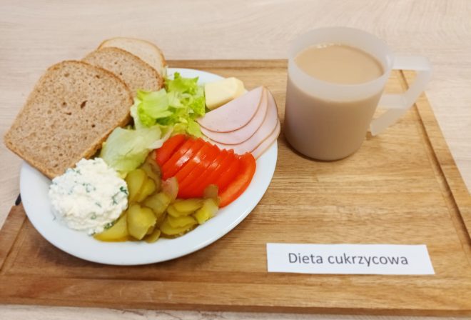 Zestaw śniadaniowy w diecie cukrzycowej, serwowany na białym talerzu. Składniki: trzy kromki chleba razowego, liście sałaty, plastry pomidora, plasterki ogórka kiszonego, serek twarogowy z ziołami, plasterki chudej wędliny drobiowej i porcja masła. Do picia kawa z mlekiem w przezroczystym kubku. Posiłek o kontrolowanej zawartości węglowodanów, urozmaicony i bogaty w błonnik