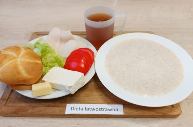 Śniadanie w diecie łatwostrawnej na drewnianej tacy. Na talerzu bułka pszenna, kostka białego twarogu, plaster masła, kilka plastrów drobiowej wędliny, liście sałaty oraz plasterki pomidora. Obok talerz z mleczną kaszą manną i kubek z herbatą ziołową. Kompozycja lekkostrawna i delikatna, odpowiednia dla osób z problemami trawiennymi