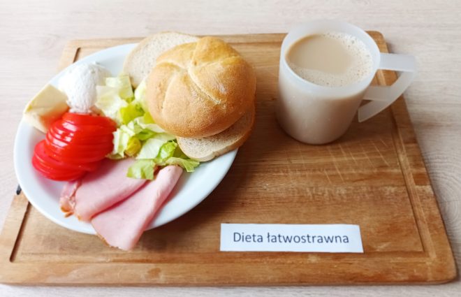 Zestaw śniadaniowy na diecie łatwostrawnej, podany na jasnej tacy. Na białym talerzu znajdują się: bułka kajzerka, dwa plastry chudej wędliny, cienkie plasterki pomidora, porcja sałaty lodowej, kulka białego twarożku oraz kawałek masła. Obok postawiono biały kubek z napojem o jasnobeżowym kolorze, przypominającym mleko z kawą zbożową. Na tacy umieszczony jest podpis „Dieta łatwostrawna”