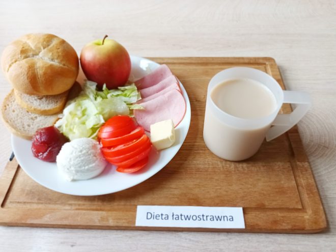 Na jasnym, drewnianym stoliku znajduje się taca z opisem „Dieta łatwostrawna”. Na niej ustawiono biały talerz z estetycznie ułożonym śniadaniem. Po lewej stronie talerza widoczne są dwie kromki jasnego pieczywa oraz bułka typu kajzerka. W centrum znajduje się sałata lodowa, kilka plastrów pomidora, plasterki gotowanej szynki oraz kostka masła. Obok umieszczono porcję białego twarożku, porcję galaretki owocowej oraz całe czerwono-żółte jabłko. Całości dopełnia biały kubek z jasnym napojem – najprawdopodobniej mlekiem lub kawą z mlekiem, stojący po prawej stronie talerza. Kompozycja posiłku jest kolorowa, lekkostrawna i zbilansowana
