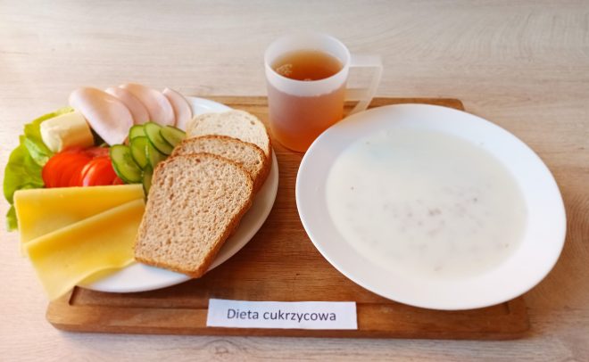 Śniadanie w diecie cukrzycowej podane na drewnianej tacy. Na talerzu umieszczono trzy kromki chleba pełnoziarnistego, plasterki chudej wędliny drobiowej, żółty ser, masło, a także świeże warzywa: plasterki ogórka, pomidora oraz sałata. Obok znajduje się biały talerz z mlekiem i drobną kaszą – prawdopodobnie zupą mleczną z płatkami zbożowymi. Do posiłku dołączono napój w przezroczystym kubku z uchwytem – najprawdopodobniej herbata. Poniżej widnieje etykieta „Dieta cukrzycowa”