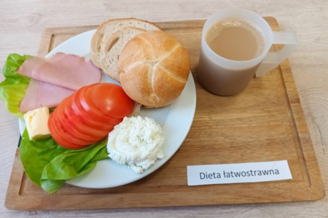 Taca z zestawem śniadaniowym przeznaczonym dla osób na diecie łatwostrawnej. Na dużym płaskim talerzu ułożono świeżą bułkę kajzerkę oraz dwa kromki jasnego chleba. Obok pieczywa znajduje się porcja białego twarożku ze szczypiorkiem, kilka plasterków pomidora, dwa cienkie plastry chudej wędliny oraz listek sałaty. Dodatkowo widoczna jest niewielka kostka masła. Po prawej stronie tacy stoi przezroczysty kubek z kawą z mlekiem. Na drewnianej podstawce znajduje się etykieta z napisem „Dieta łatwostrawna”
