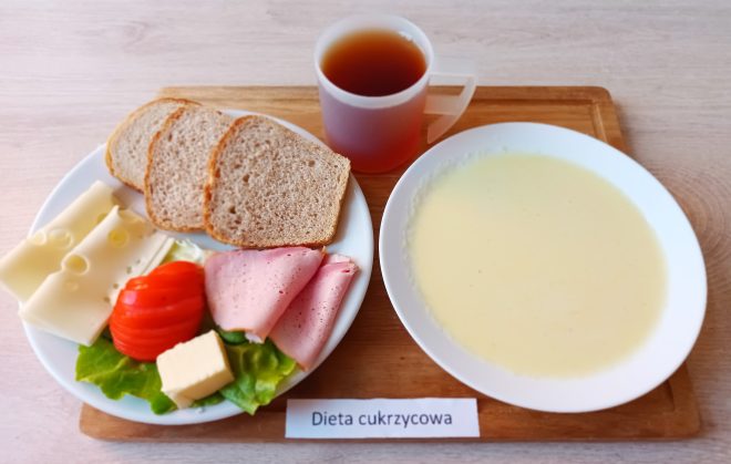 Na fotografii przedstawiono śniadanie dostosowane do potrzeb osoby na diecie cukrzycowej, ułożone na jasnej tacy z drewnianym spodem i etykietą „Dieta cukrzycowa”. Po lewej stronie znajduje się talerz z trzema kromkami pieczywa razowego, trzema plasterkami wędliny drobiowej, kilkoma plasterkami sera żółtego typu ementaler oraz świeżymi warzywami – plasterkami pomidora i liśćmi sałaty. Na talerzu ułożono także kostkę białego twarogu. Po prawej stronie znajduje się talerz z jasnym kremem – prawdopodobnie zupa mleczna lub kisiel mleczny. Nad posiłkiem stoi przezroczysty kubek z herbatą bez cukru. Kompozycja śniadania sugeruje dobrze zbilansowany, łagodny posiłek przeznaczony dla osób z cukrzycą, z uwzględnieniem niskiego indeksu glikemicznego