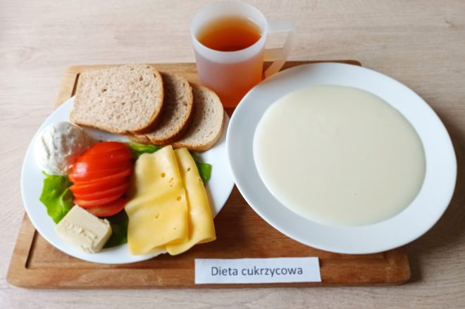 Zestaw śniadaniowy oznaczony jako „Dieta cukrzycowa”, podany na drewnianej tacy. Na jednym talerzu znajduje się kilka kromek pieczywa żytniego, plasterki pomidora i żółtego sera, liść sałaty, kawałek białego twarogu oraz kostka białego sera typu feta. Na drugim talerzu – gładka zupa mleczna o jasnym kolorze. Obok postawiono przezroczysty kubek z ciemniejszym napojem, najprawdopodobniej herbatą bez cukru