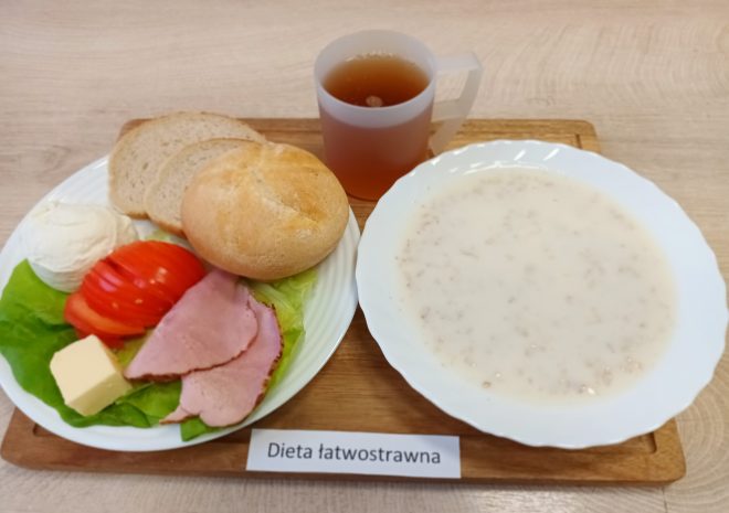 Taca z posiłkiem oznaczonym jako dieta łatwostrawna ustawiona na jasnym, drewnopodobnym blacie. Na tacy znajdują się trzy elementy: Główny talerz – Na białym talerzu ułożono porcję zupy mlecznej z drobną kaszą, przypominającą mannę lub płatki owsiane. Zupa ma kremową, mleczną konsystencję z wyraźnie widocznymi drobinkami kaszy. Talerz śniadaniowy – Zawiera jasną bułkę kajzerkę oraz dwa plasterki chleba pszennego. Obok pieczywa ułożono: dwa cienkie plasterki gotowanej szynki, plaster sera żółtego, liść sałaty zielonej, plasterki pomidora, kopiastą porcję białego twarożku. Napój – W przezroczystym kubku z matowego plastiku znajduje się napar – prawdopodobnie słaba herbata ziołowa lub owocowa, o jasnobrązowym kolorze. Pod talerzami umieszczona jest biała karteczka z nadrukiem: "Dieta łatwostrawna"