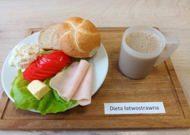 Śniadanie serwowane w ramach diety łatwostrawnej, podane na drewnianej tacy. Na talerzu widoczne są dwa plasterki pieczywa (kromka chleba i bułka pszenna typu kajzerka), liść sałaty z trzema składnikami: porcją twarogu, plastrami świeżego pomidora oraz dwoma cienkimi plasterkami wędliny drobiowej. Obok znajduje się niewielka kostka masła. Całość uzupełnia plastikowy kubek z jasnobrązowym napojem – prawdopodobnie kawa zbożowa lub kawa z mlekiem, o delikatnej konsystencji. Na tacy umieszczono etykietę z napisem „Dieta łatwostrawna”