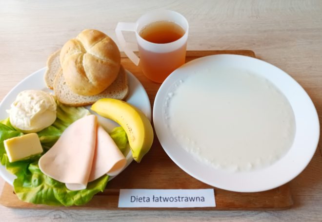 Zestaw śniadaniowy na diecie łatwostrawnej, podany na jasnym drewnianym stoliku. Na białym talerzu widoczna bułka kajzerka oraz dwa plasterki chleba pszennego. Obok ułożono plaster żółtego sera, dwa plasterki wędliny drobiowej, liść sałaty, połówkę banana i kulkę białego twarożku. Na osobnym talerzu znajduje się zupa mleczna lub kaszka mleczna. Całość uzupełnia przezroczysty kubek z herbatą. Z przodu etykieta z napisem „Dieta łatwostrawna”