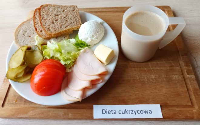Taca z posiłkiem śniadaniowym opisana etykietą „Dieta cukrzycowa”. Na talerzu znajduje się starannie ułożony zestaw: trzy kromki chleba razowego, plasterki chudej wędliny z indyka lub kurczaka, twarożek w formie kulki, kostka masła, kilka plasterków ogórka kiszonego, sałata lodowa oraz pokrojony pomidor. Danie uzupełnia kubek jasnej kawy z mlekiem lub zbożowej kawy z mlekiem, w przezroczystym, plastikowym kubku z uchem. Całość ustawiona jest na drewnianej tacy, na jasnym stole